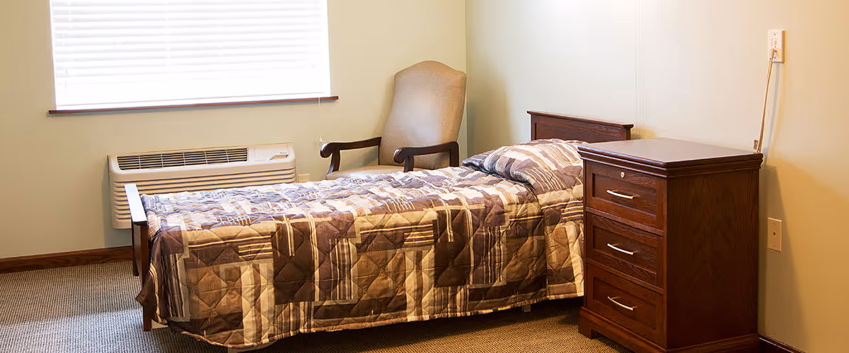 A simple bedroom with a single bed covered in a patterned quilt, a wooden nightstand with three drawers, a beige upholstered armchair, and a window with blinds above an air conditioning unit.