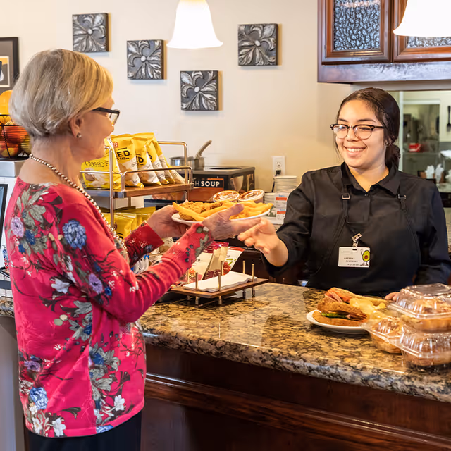 A smiling staff member behind a service counter hands a plate of food to an older woman in a dining area.