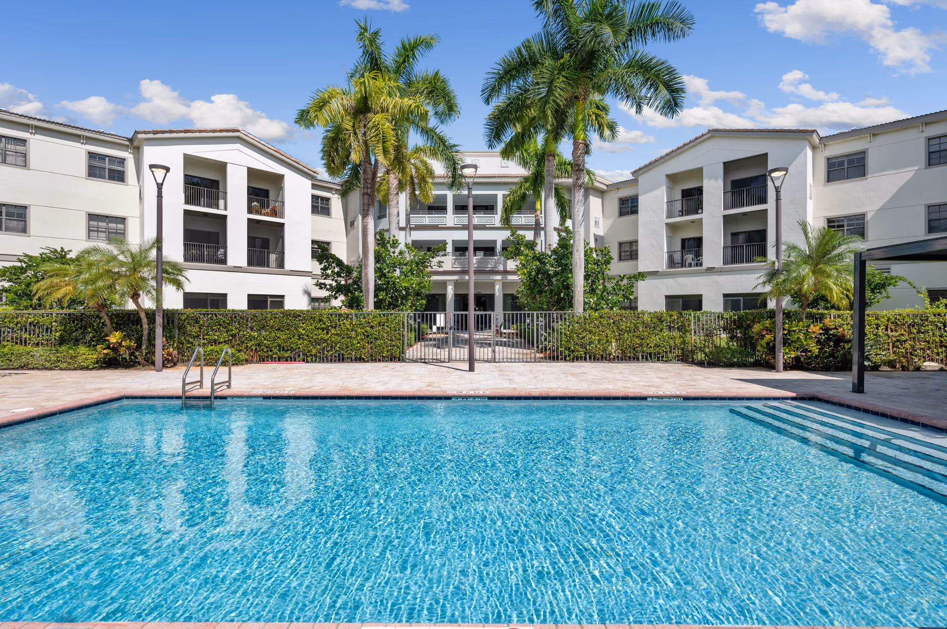 Outdoor swimming pool in a courtyard in front of a multi-story residential building with palm trees and balconies.