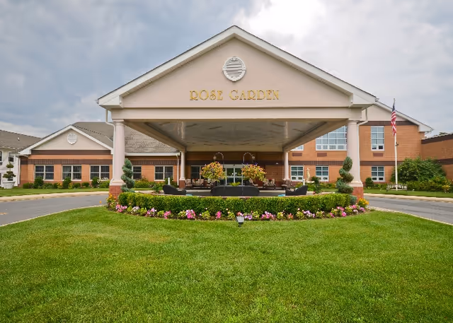 Front exterior view of Rose Garden Nursing and Rehabilitation Center showing a covered entrance with columns, a circular driveway, well-maintained green lawn, flower beds, and an American flag on a flagpole to the right.