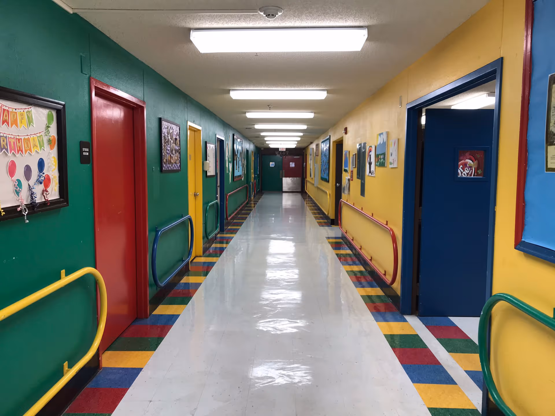 A brightly colored hallway in a children's care center with green, yellow, and blue walls, multicolored floor tiles along the edges, handrails on both sides, and various framed artworks and decorations on the walls. Several doors are visible along the hallway, including a red door labeled 'Stock Room'.