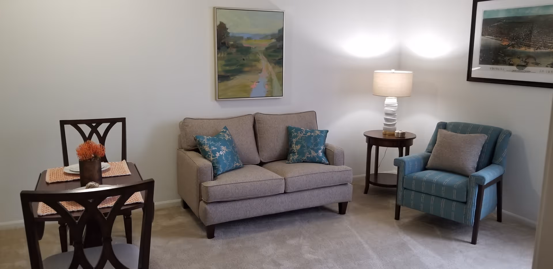 A cozy living area featuring a beige loveseat with two blue patterned throw pillows, a blue armchair with a gray pillow, a small round wooden side table with a white lamp, and a small dining table with two chairs and a centerpiece. The walls are decorated with two framed paintings.