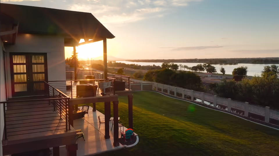 Two-story deck and patio attached to a house overlooking a grassy yard and a river at sunset.