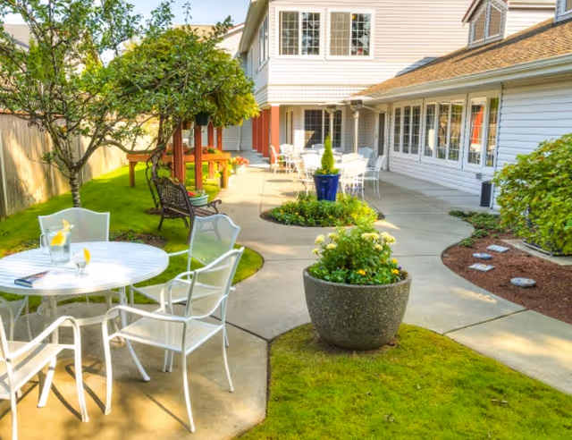 Outdoor patio area at The Terrace at Beverly Lake by Cogir featuring white metal tables and chairs, a large potted plant with flowers, a tree, and a pathway leading to a building with multiple windows and doors.