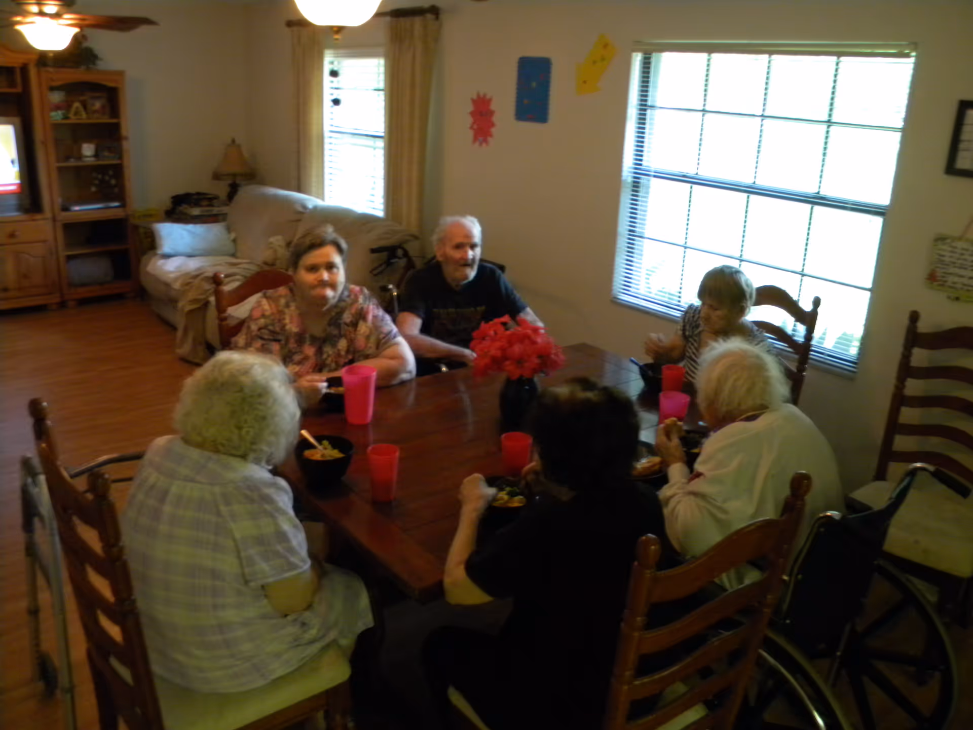 A group of elderly people sitting around a wooden dining table in a well-lit room with large windows. They are eating from bowls and drinking from red plastic cups. The room has wooden floors, a beige couch, a wooden cabinet with a TV, and some wall decorations.