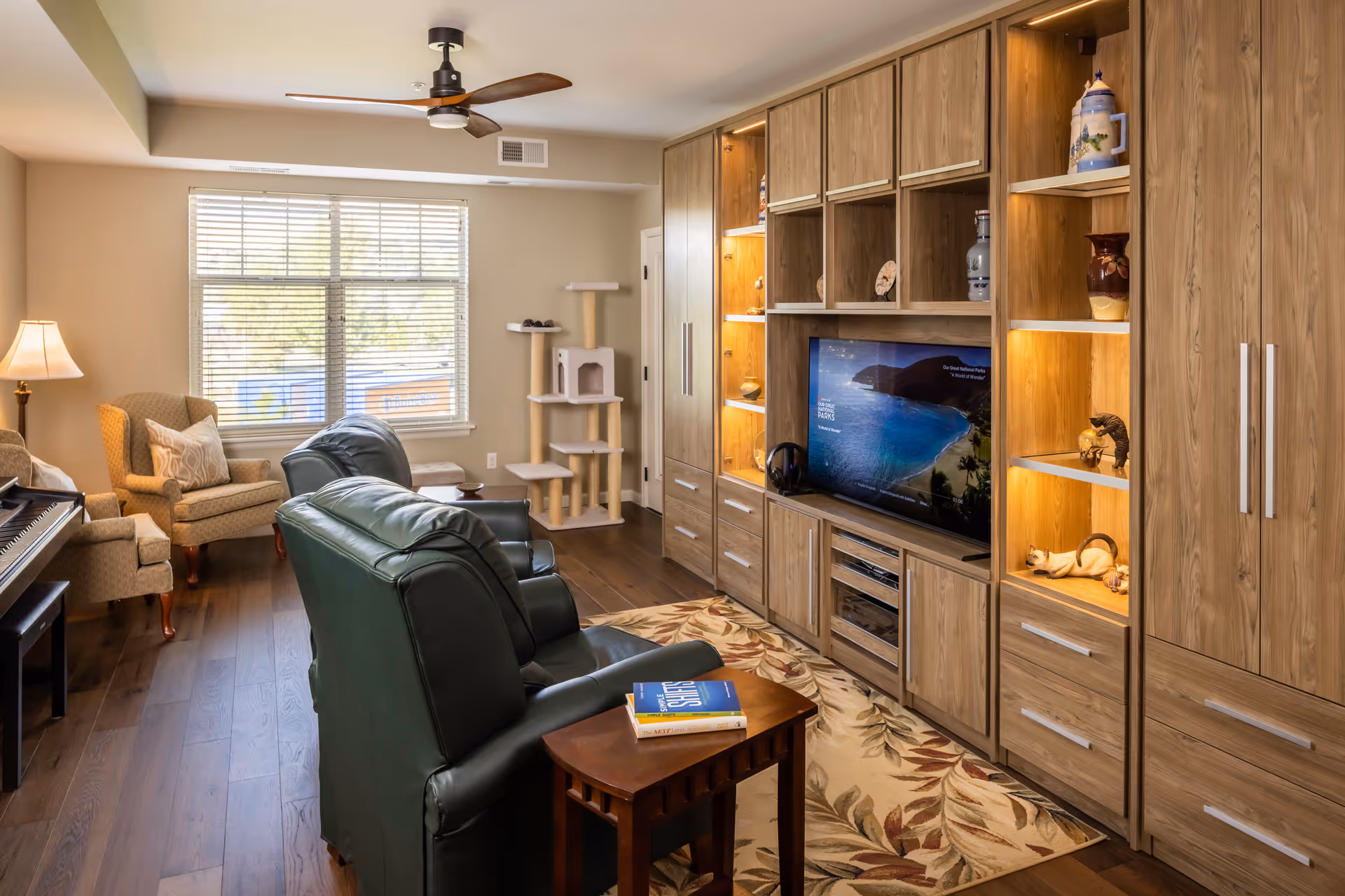 A cozy living room with two green leather recliners facing a large wooden entertainment center with a flat-screen TV displaying a beach scene. The entertainment center has shelves with decorative items and cabinets. There is a beige armchair with a pillow near a window with blinds, a floor lamp, a cat tree, and a piano on the left side. The room has wooden flooring, a ceiling fan, and a floral area rug.