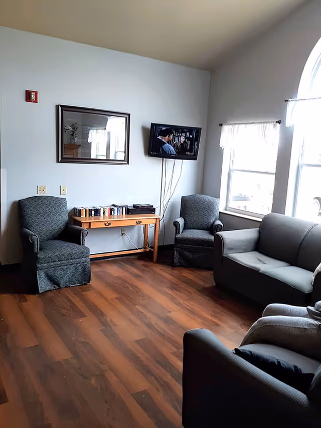 A cozy living room area with wooden flooring, two upholstered armchairs, a gray sofa, a wooden table with books and a DVD player, a wall-mounted TV showing a scene from a movie, a mirror on the wall, and windows with white curtains letting in natural light.