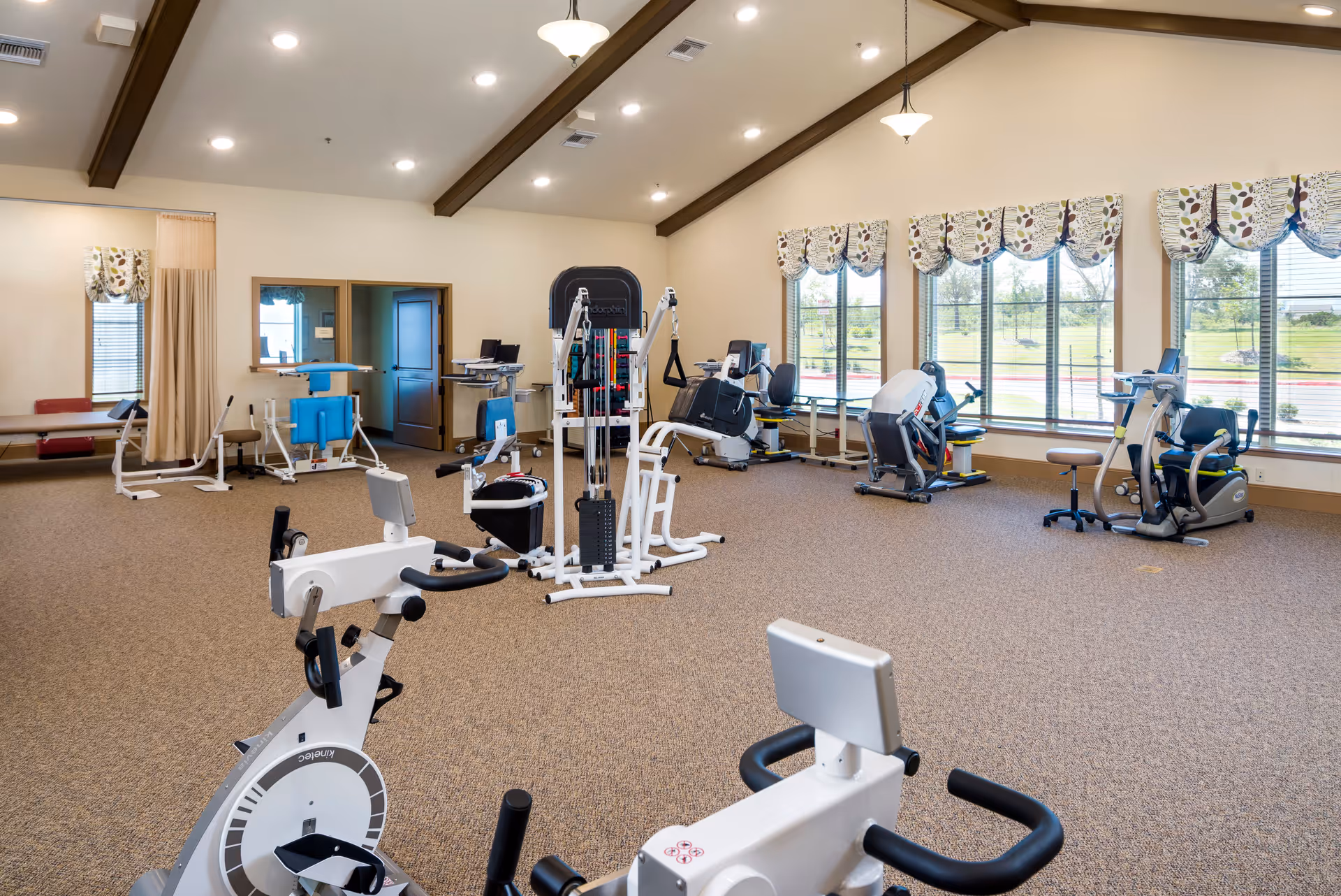 A spacious exercise room with various fitness equipment including stationary bikes, weight machines, and other exercise apparatus. The room has large windows with patterned valances allowing natural light to brighten the space. The ceiling features exposed wooden beams and recessed lighting.