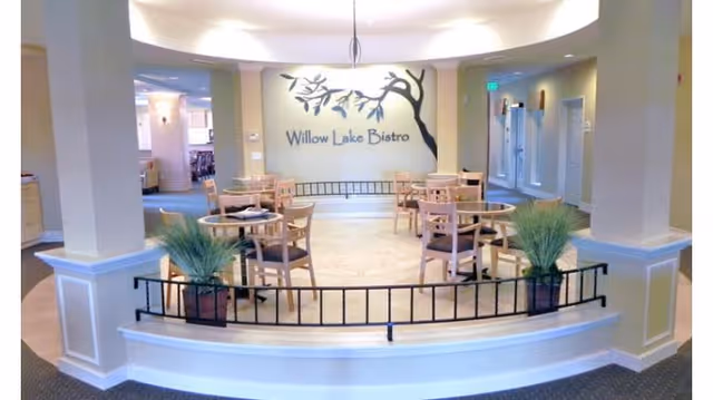 Interior view of a dining area named Willow Lake Bistro with several round wooden tables and chairs arranged neatly. The space is well-lit with soft lighting and decorated with potted plants and a wall mural featuring a tree branch and the bistro name.