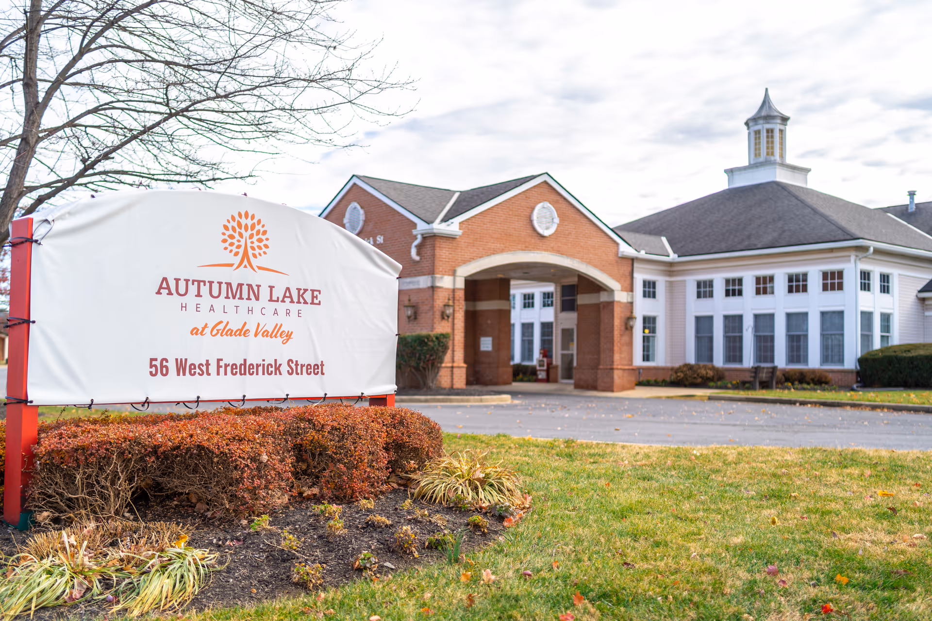 Exterior view of Autumn Lake Healthcare at Glade Valley building with a large sign in the foreground displaying the facility name and address, 56 West Frederick Street. The building features a brick entrance with an archway and white-framed windows, surrounded by grass and landscaping.