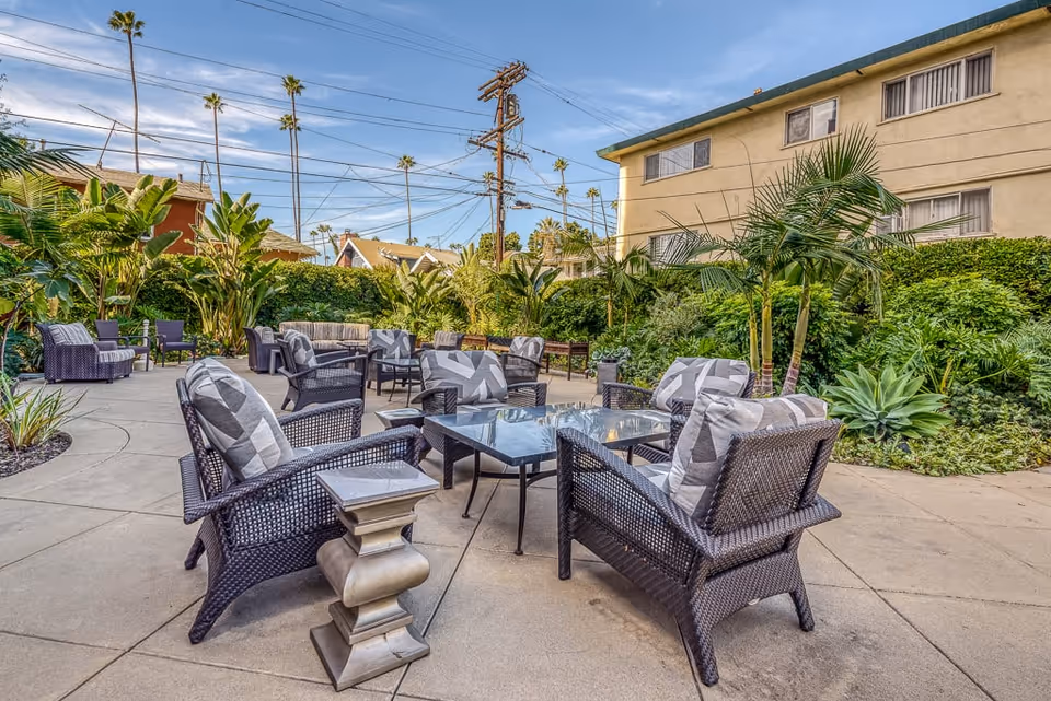 Outdoor patio area with multiple wicker chairs and glass-top tables arranged on a concrete surface, surrounded by lush green plants and palm trees, with a residential building and clear blue sky in the background.