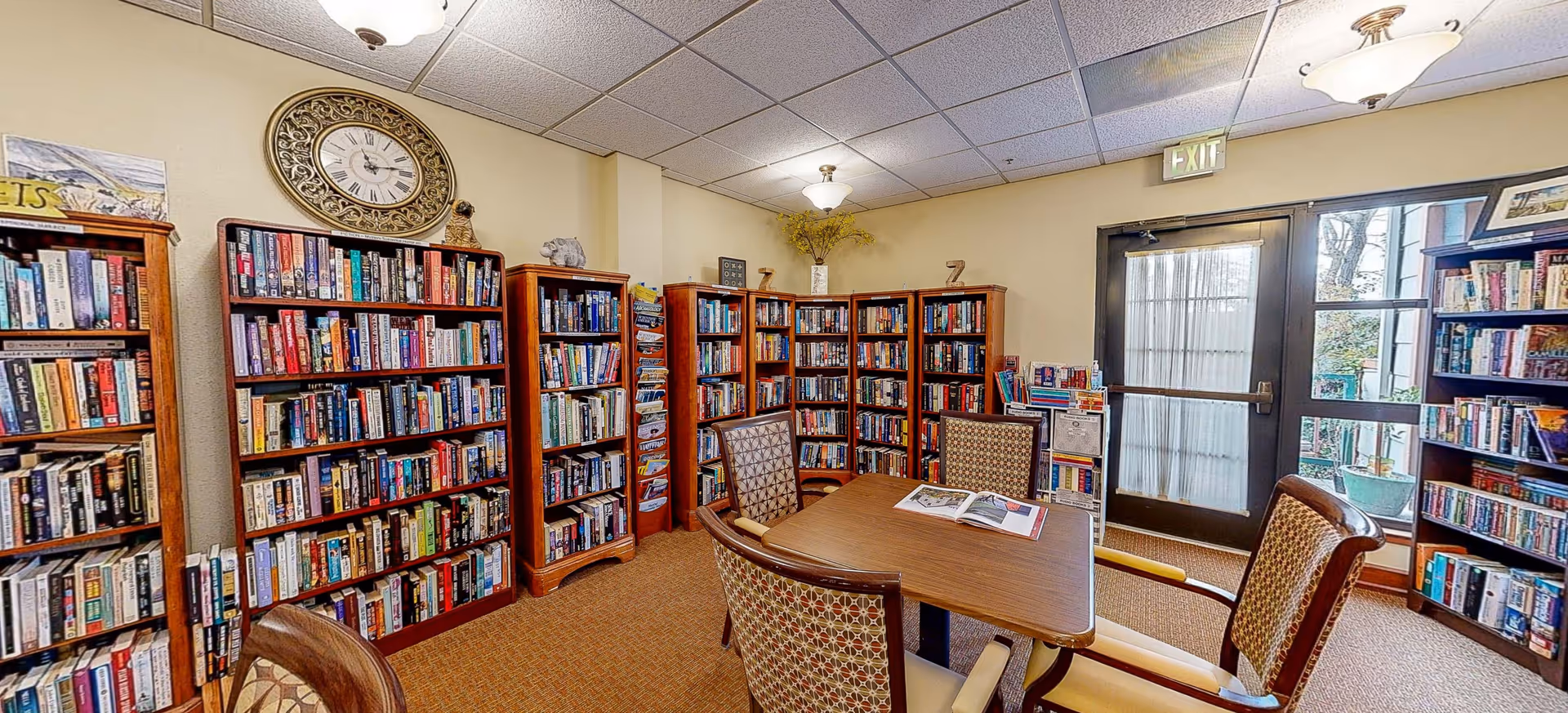 Cozy reading room with bookshelves lining the walls, a central table and chairs, and a glass exit door.
