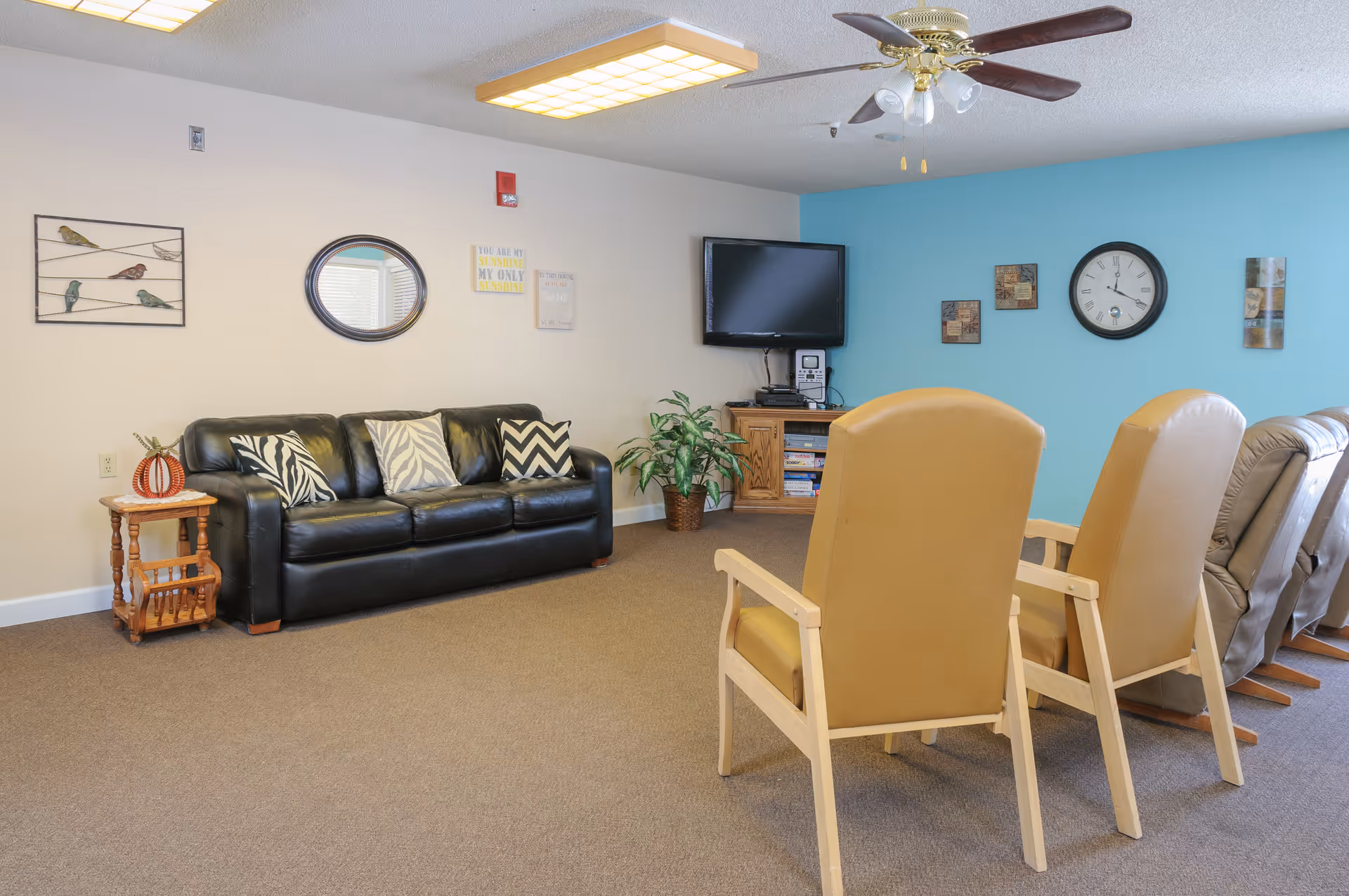 A cozy living room area with a black leather couch adorned with patterned pillows, two beige armchairs, and two recliners. The walls are decorated with a round mirror, bird artwork, a clock, and other framed pieces. A flat-screen TV is mounted on a blue accent wall above a wooden cabinet with books and a stereo system. A ceiling fan with lights is overhead.