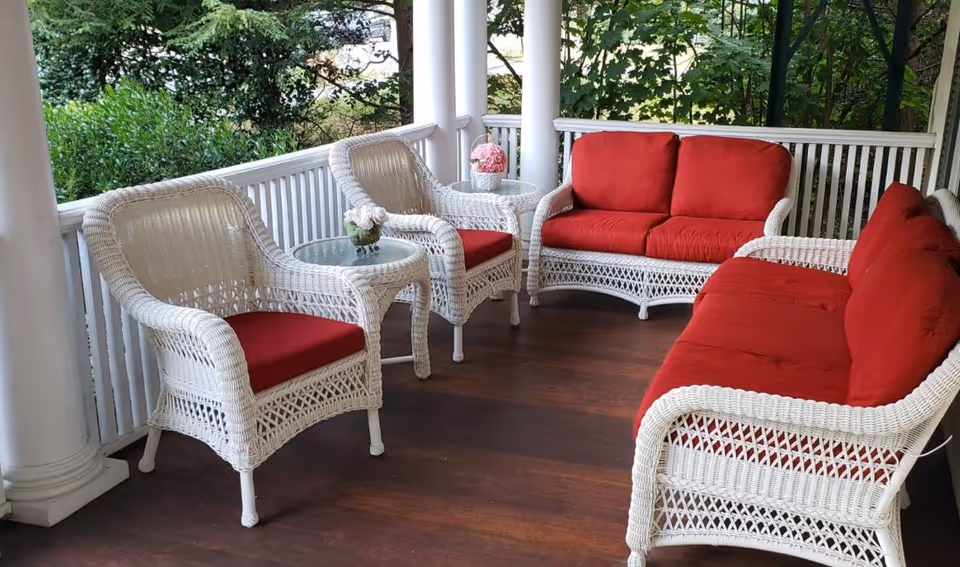 A porch area with white wicker furniture including two armchairs with red cushions, two glass-top side tables with flower arrangements, and two sofas with red cushions. The porch has white railings and columns, with greenery visible outside.