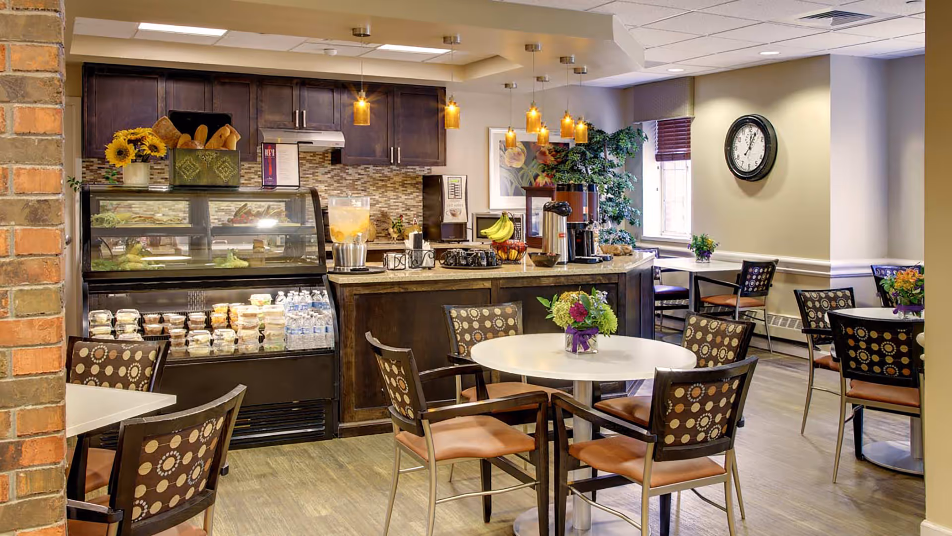 A cozy dining area in a senior living facility featuring round tables with floral centerpieces and patterned chairs. In the background, there is a counter with a coffee machine, fruit, and a display case containing bottled water and packaged food items. Warm pendant lights hang from the ceiling, and a large clock is mounted on the wall near a window with blinds.