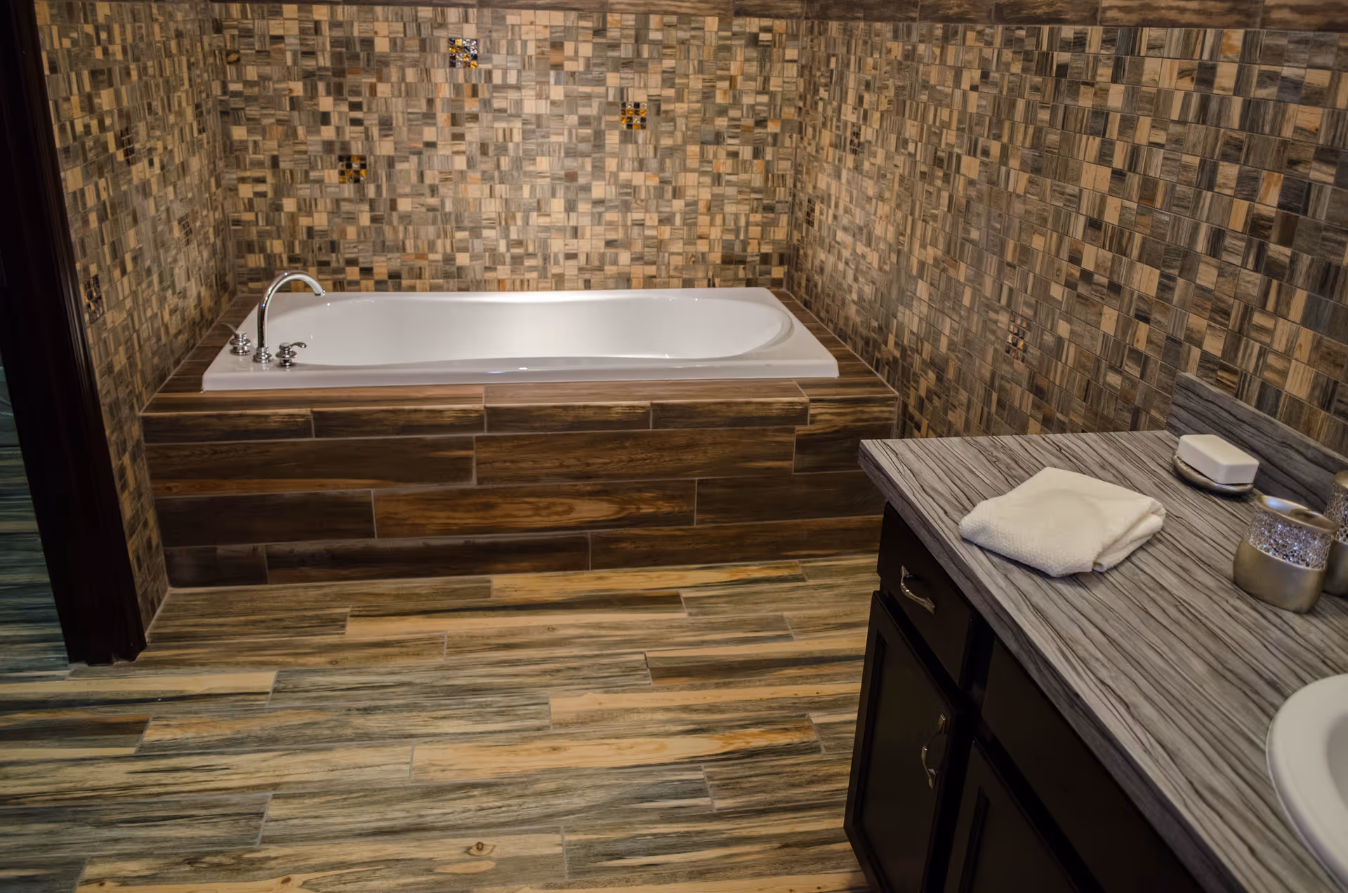 A bathroom with a built-in bathtub surrounded by wood-like tiles and mosaic tiled walls. The floor has wood-patterned tiles. A countertop with a folded white towel, soap dish, and decorative items is visible on the right side.