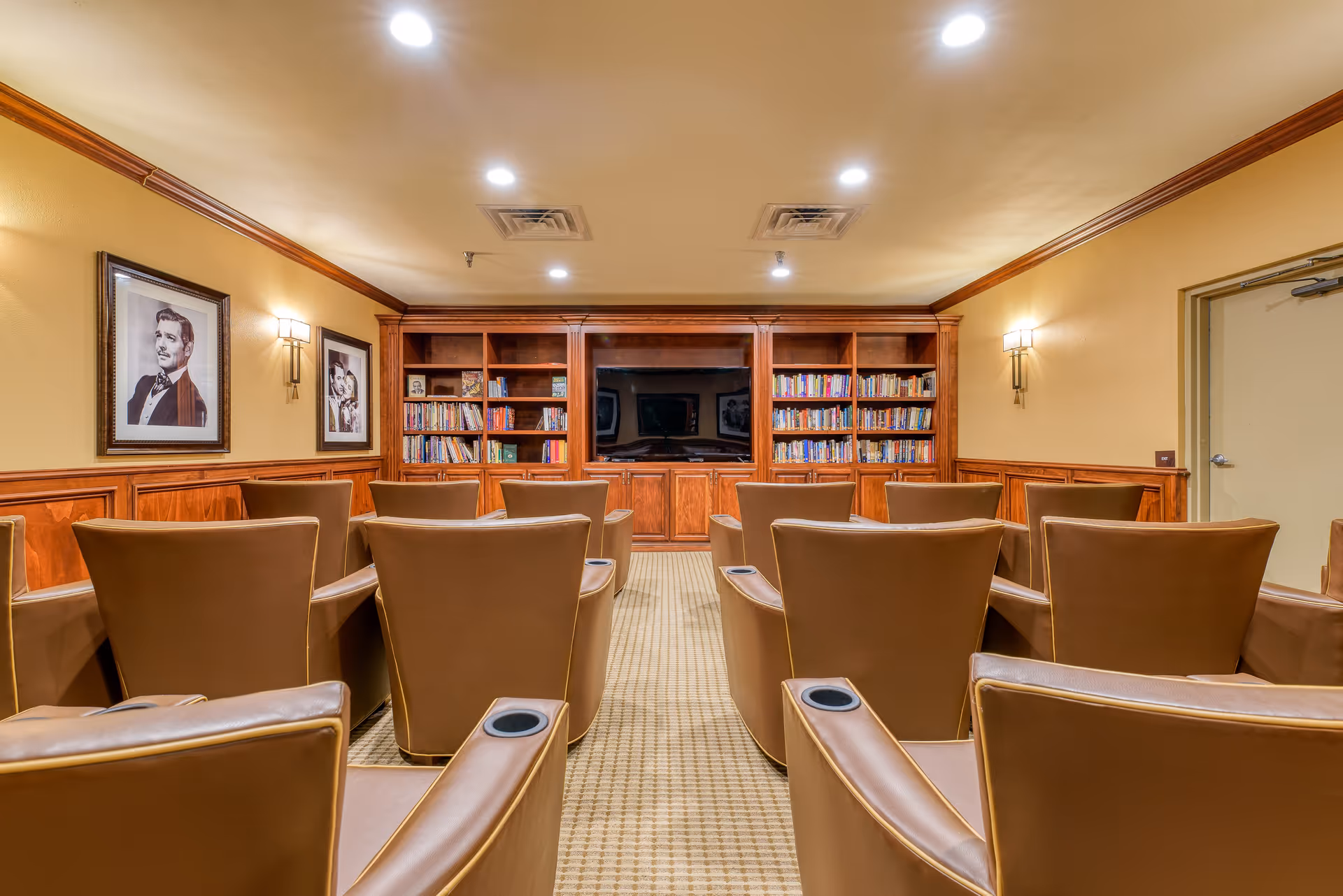 A cozy media room with rows of brown leather chairs facing a large flat-screen TV mounted on a wooden bookshelf filled with books. The room has warm yellow walls, wood paneling, framed black and white portraits on the left wall, and recessed ceiling lights.