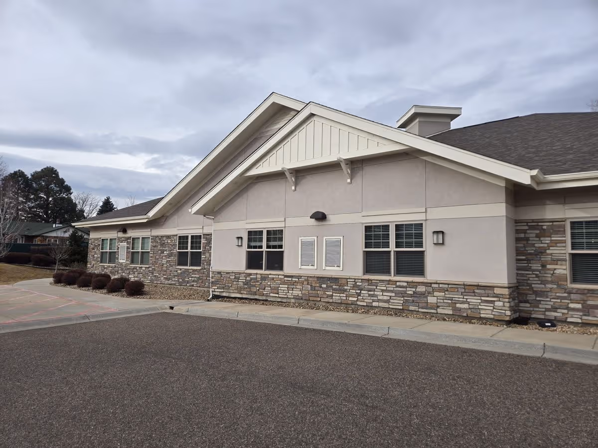 Exterior front view of a single-story memory care building with stone veneer, windows, and a peaked roof under a cloudy sky.