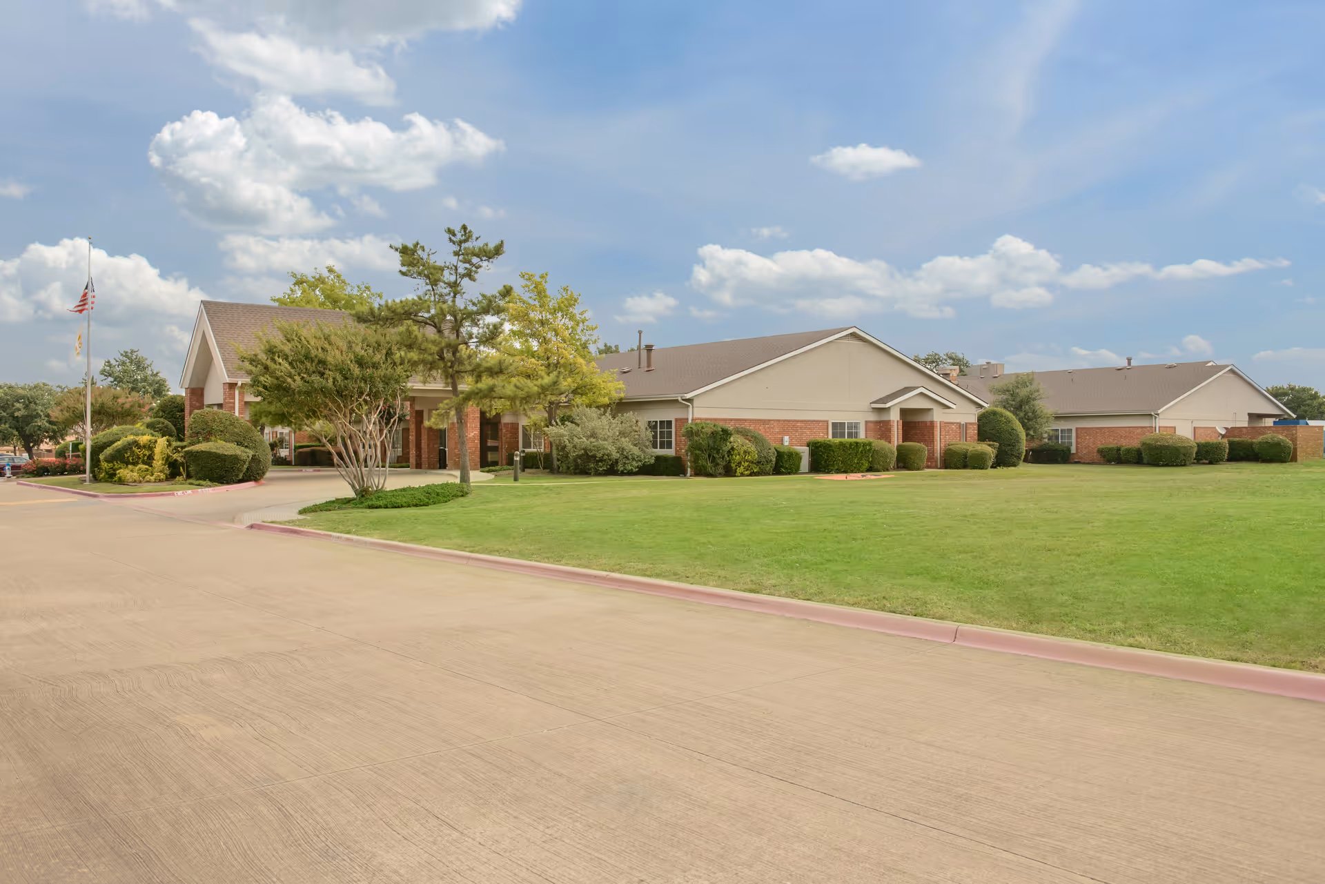 Exterior view of a single-story senior living facility building with a brick and beige facade, surrounded by green grass, bushes, and trees under a partly cloudy sky.