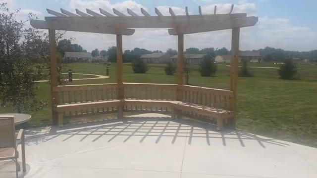 A wooden pergola with built-in benches on a concrete patio overlooking a large grassy area with trees and houses in the distance under a partly cloudy sky.