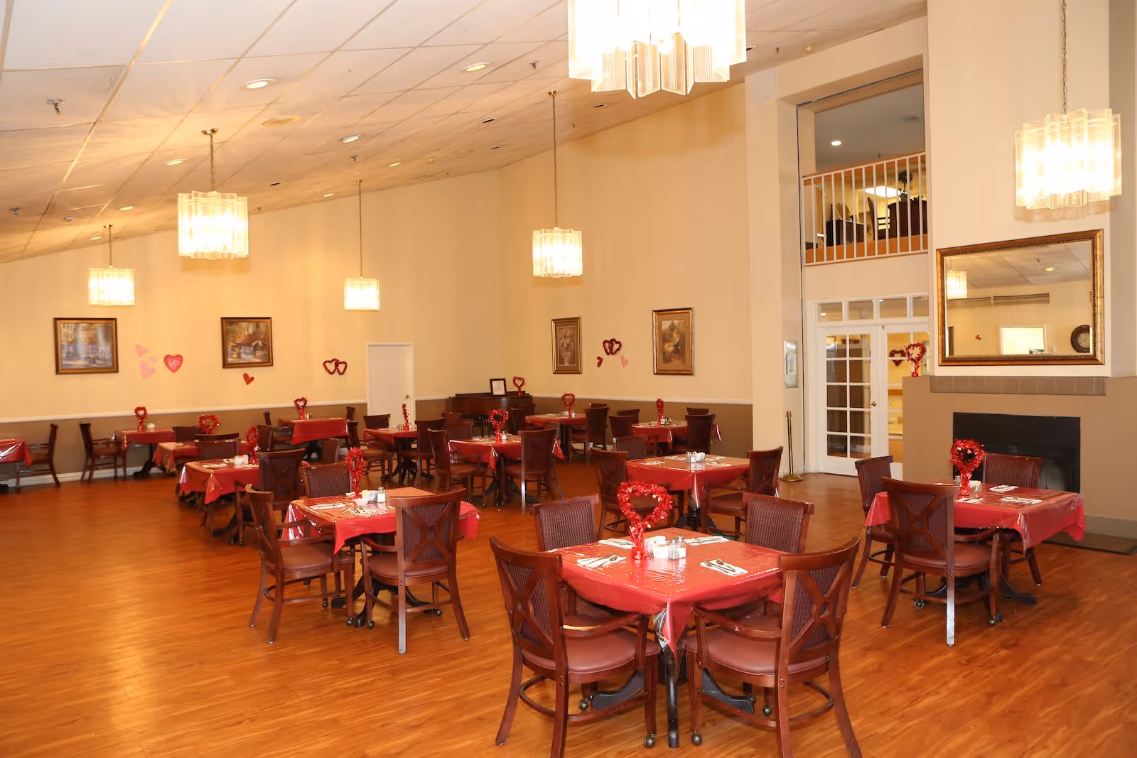 A spacious dining room with multiple tables covered in red tablecloths and decorated with heart-shaped centerpieces. The room has wooden flooring, beige walls, and several hanging light fixtures. There are framed paintings on the walls and a large mirror above a fireplace. The room appears well-lit and ready for a meal service.