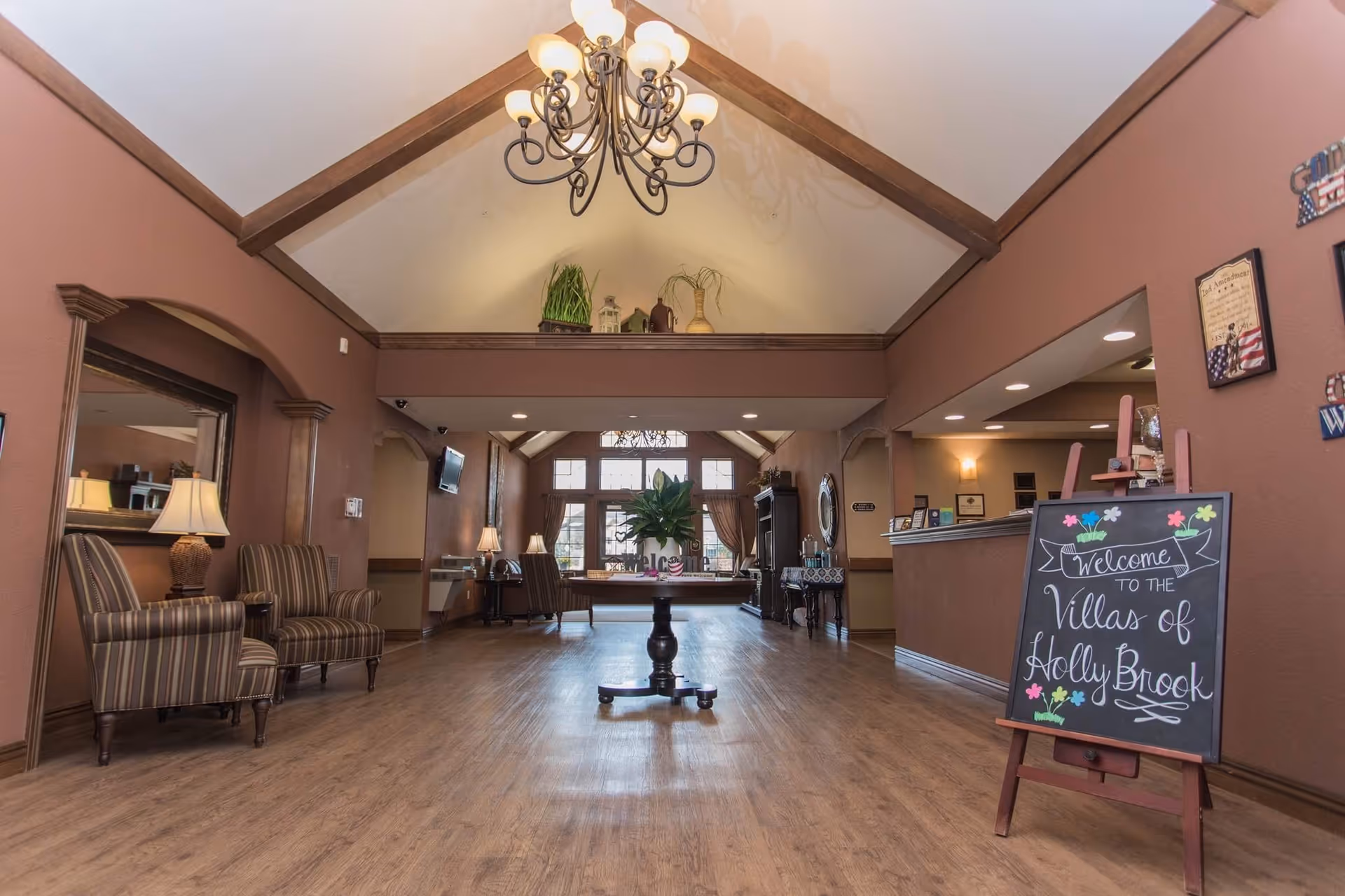 Interior view of a senior living facility lobby with a high vaulted ceiling and chandelier. The room features wooden flooring, two striped armchairs with a lamp on the left, a central round table with a plant, and a reception desk on the right. A chalkboard sign near the reception desk reads 'Welcome to the Villas of Holly Brook'. Large windows with curtains are visible at the far end of the room.