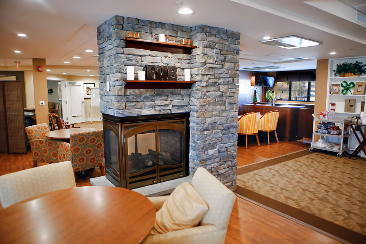 Interior view of a senior living facility lounge area featuring a stone fireplace with shelves holding candles and decorative letters spelling 'PUB'. Surrounding the fireplace are round wooden tables with cushioned chairs. In the background, there is a bar area with two striped chairs, a TV mounted on the wall, and shelves with plants and framed pictures.
