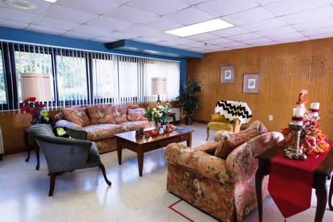 Cozy common room with floral sofas and armchairs arranged around a wooden coffee table beneath large windows with vertical blinds.
