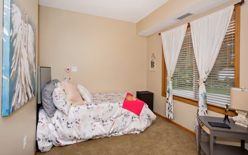 Cozy single bedroom with a floral-covered bed, pillows and a bright pink throw, a window with white curtains, bedside table and wall art.