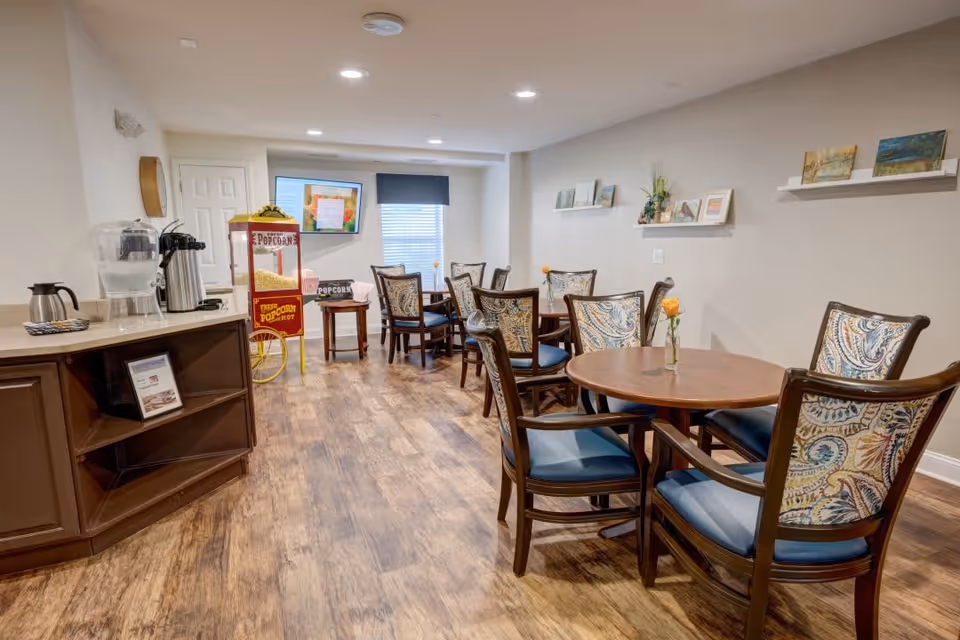 A cozy dining area with several round wooden tables and cushioned chairs featuring a paisley pattern. On the left side, there is a counter with a water dispenser, coffee pot, and other beverage supplies. In the background, a vintage-style popcorn machine and a small table with a popcorn sign are visible. The walls are decorated with small shelves holding framed pictures and plants. The room has wood flooring and is well-lit with recessed ceiling lights.