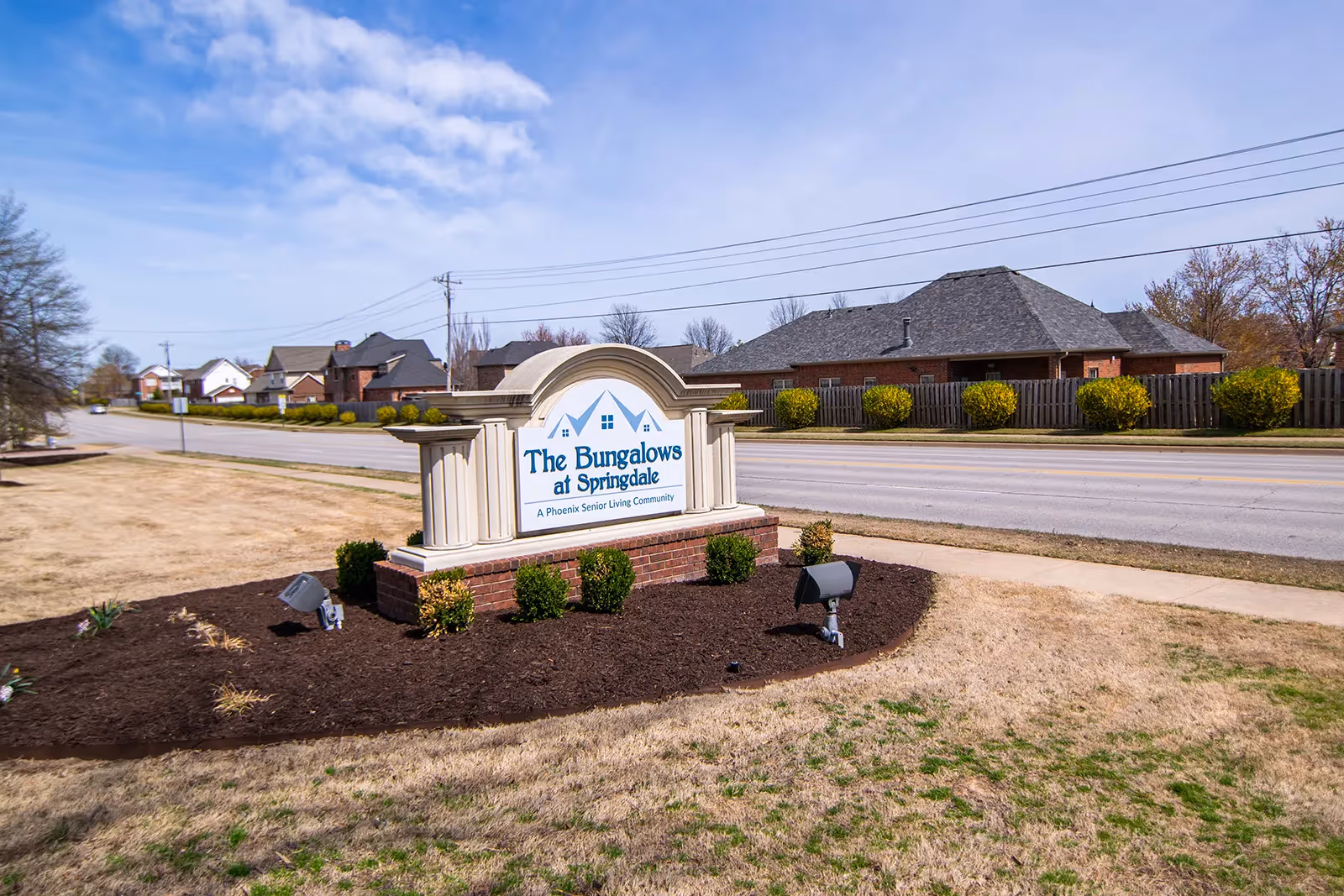 Outdoor view of the entrance sign for The Bungalows at Springdale, a Phoenix senior living community, with a road and residential houses in the background under a partly cloudy sky.