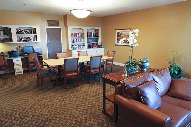 Community lounge with a leather sofa, a long table and chairs, bookshelves and a small computer desk.