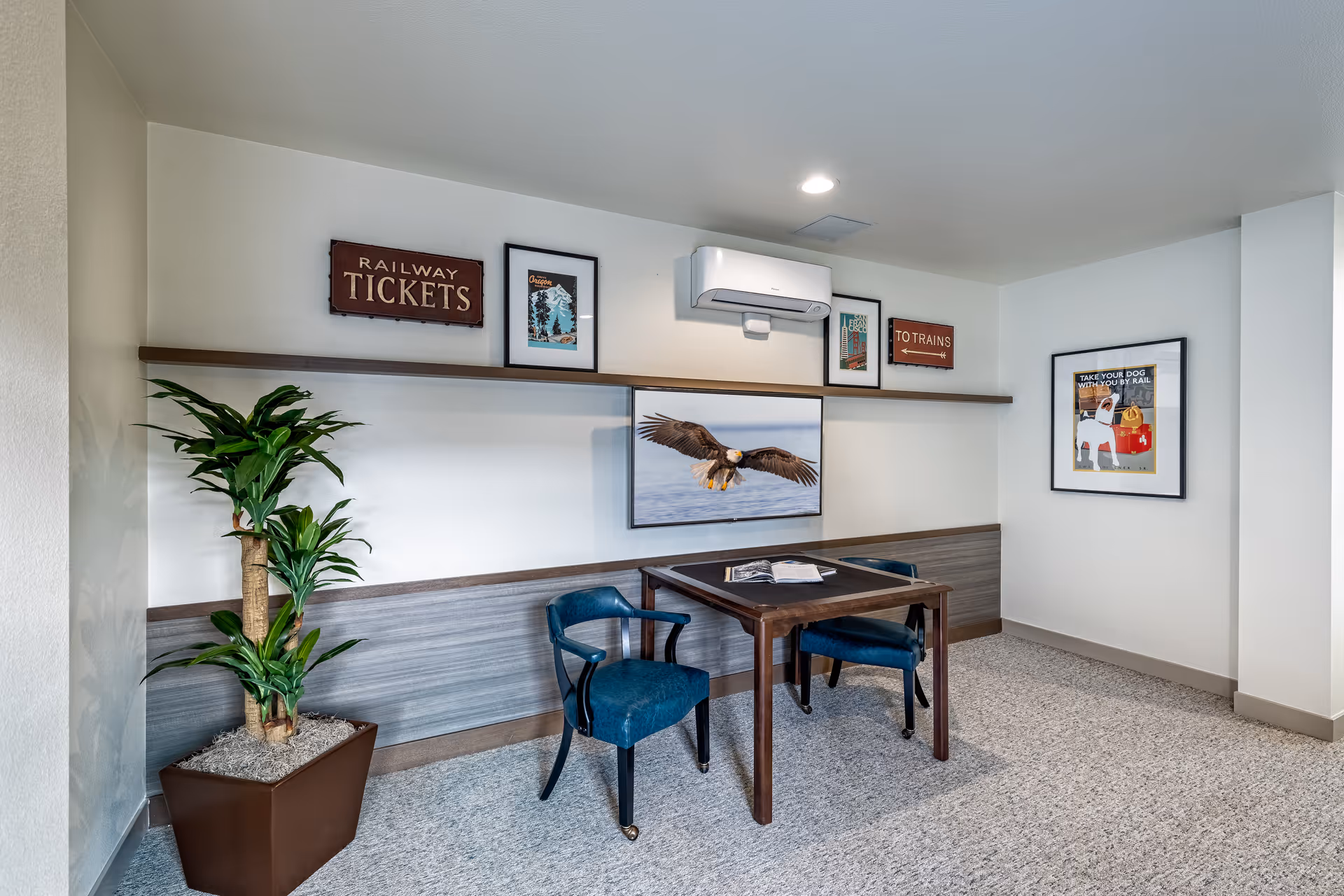 A small seating area with a wooden table and two blue upholstered chairs on a carpeted floor. The wall behind has a long wooden shelf displaying framed pictures and signs, including one that says 'RAILWAY TICKETS' and another that says 'TO TRAINS'. A framed picture of a bald eagle in flight is mounted below the shelf. A potted plant is placed on the left side of the seating area. The walls are painted white and there is a ceiling light above.