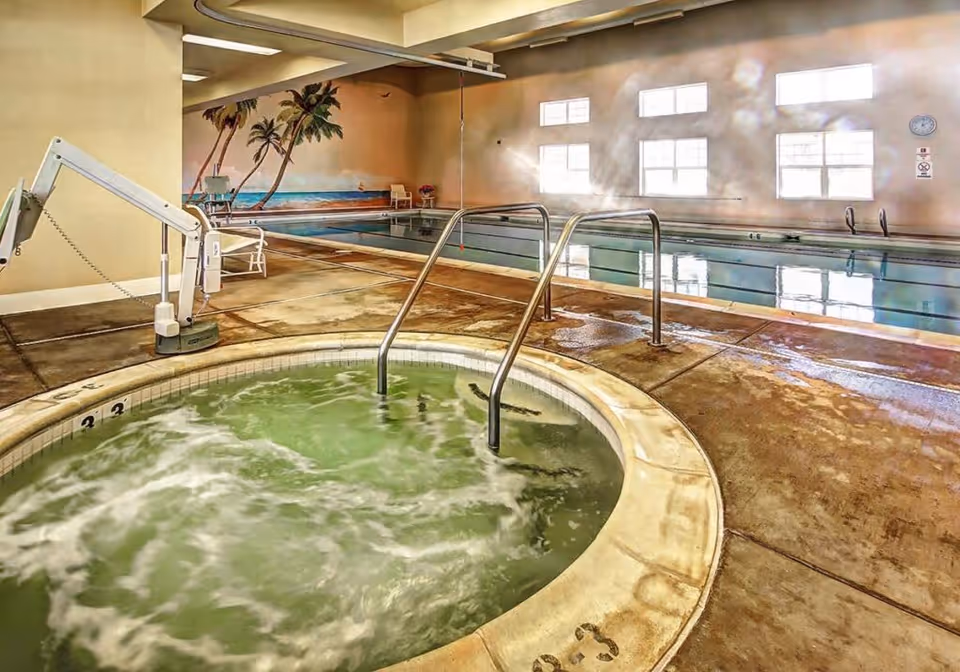 Indoor pool area with a hot tub in the foreground and a swimming pool in the background. The hot tub has bubbling water and metal handrails. The pool area features a mural of palm trees and a beach scene on the far wall. There are large windows letting in natural light and a pool lift chair for accessibility.