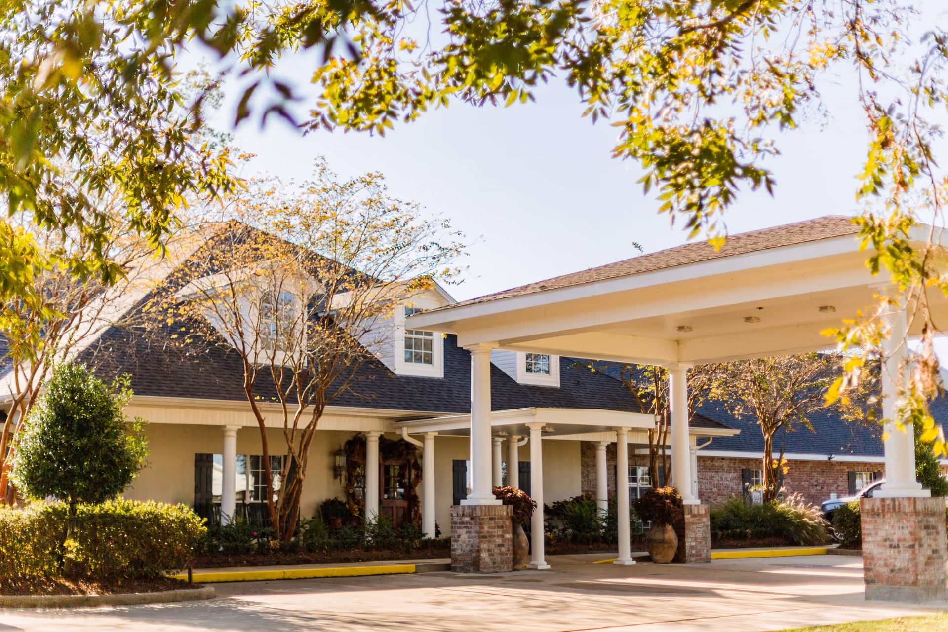 Exterior view of Oakmont Estates showing a covered entrance with white columns, brick pillars, and a sloped roof. Trees and shrubs surround the building under a clear sky.