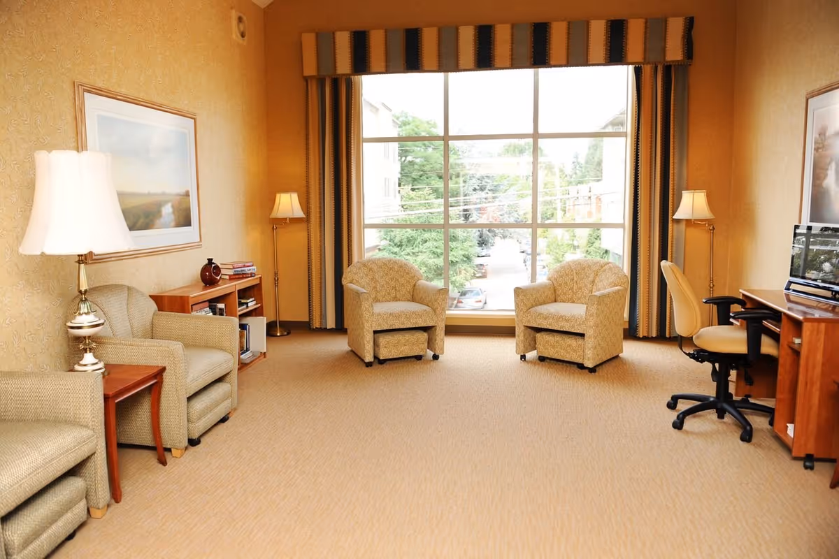 A bright and cozy living room with beige carpet and walls, featuring four upholstered armchairs arranged around a large window with striped curtains. There are two floor lamps and a table lamp providing warm lighting. A wooden desk with an office chair and a fish tank is positioned on the right side, and a small bookshelf with books and decorative items is on the left.