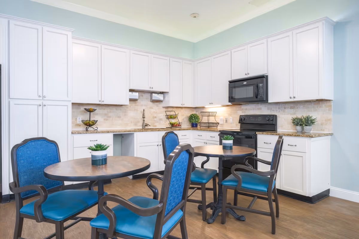 A bright kitchen area with white cabinets, a black microwave and stove, and granite countertops. There are two round wooden tables with blue cushioned chairs around them. Small potted plants are placed on the tables and countertops. The walls are painted light blue and the floor is wooden.
