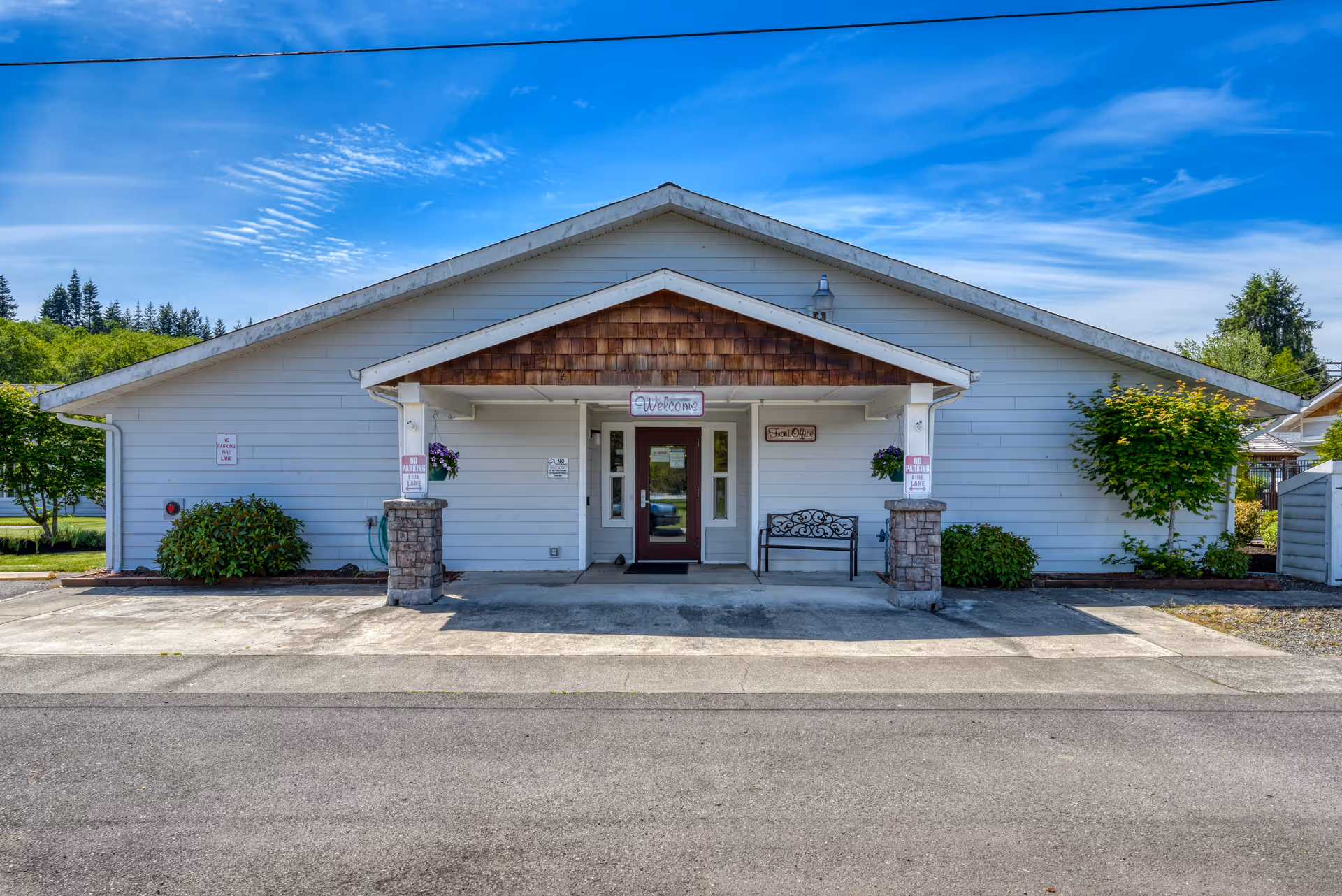 Front exterior view of a single-story building with white siding and a peaked roof. The entrance has a covered porch with two stone pillars, a bench, and hanging flower baskets. A sign above the door reads 'Welcome'. There are small bushes and trees on either side of the building, and a clear blue sky is visible in the background.