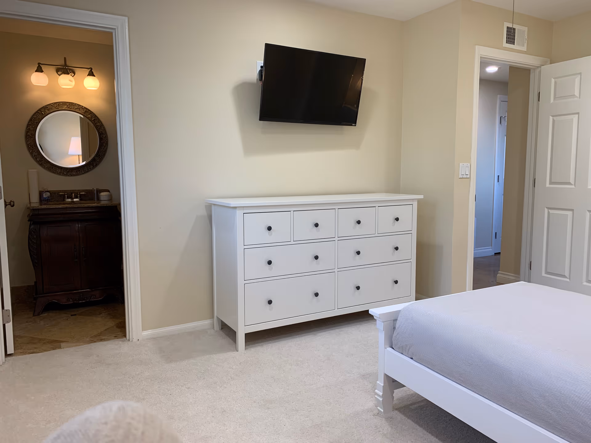 A bedroom with a white bed frame and mattress, a white dresser with multiple drawers, and a wall-mounted flat-screen TV above the dresser. To the left, there is an open door leading to a bathroom with a round mirror and a vanity with a lamp reflected in the mirror. The walls are painted beige and the floor is carpeted.