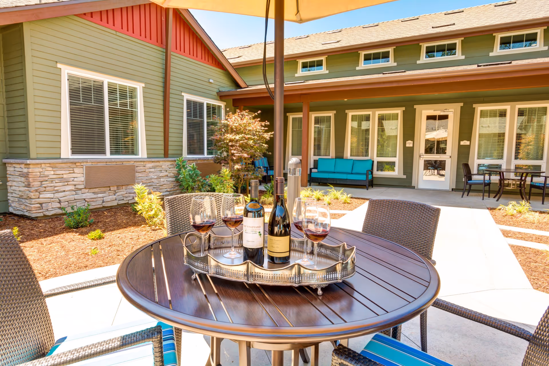 Outdoor patio area at The Vineyard at Fountaingrove Memory Care featuring a round wooden table with a tray holding two bottles of wine and four glasses of red wine. The table is surrounded by wicker chairs with blue-striped cushions. In the background, there is a green building with white-trimmed windows, a covered porch with a teal cushioned bench, and another table with chairs. The patio is shaded by a large umbrella.