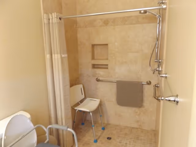 Accessible shower area with beige tiled walls and floor, a white shower chair with blue feet, a grab bar with a gray towel hanging on it, and a shower curtain on a curved rod. Part of a white toilet with armrests is visible on the left side.
