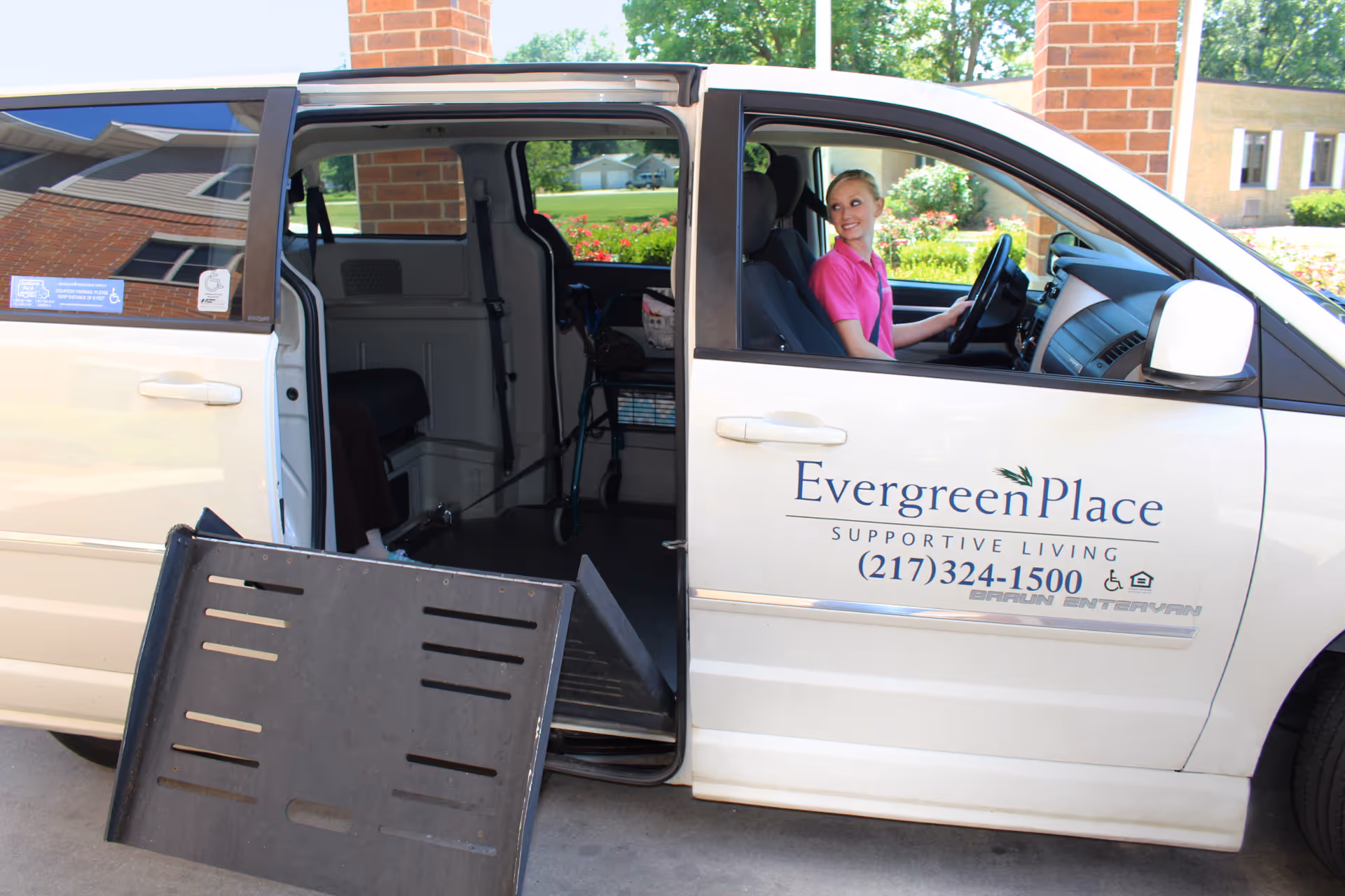 White Evergreen Place supportive living van with its sliding door open, a deployed wheelchair ramp, and a smiling driver at the wheel.