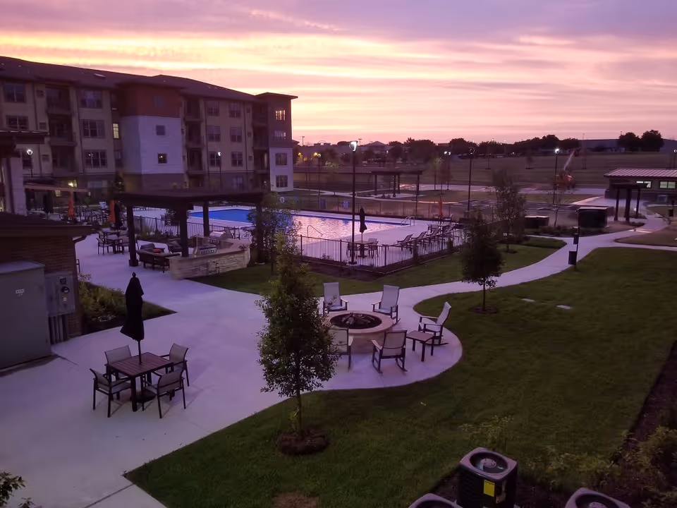 Outdoor area of a senior living facility at sunset with a swimming pool surrounded by lounge chairs and umbrellas, a fire pit with chairs around it, tables with chairs and umbrellas, paved walkways, small trees, and a multi-story residential building in the background.