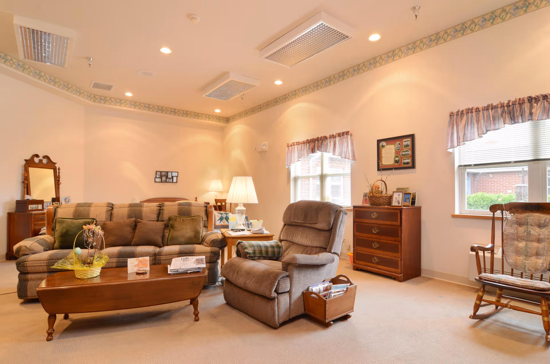 Comfortable common living room featuring a plaid sofa, recliner, coffee table, rocking chair and dresser under warm overhead lighting.