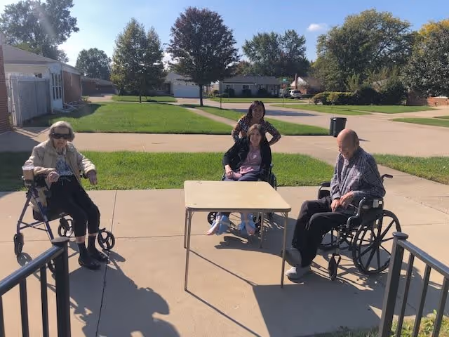 Three elderly individuals sitting outside on a paved area, two in wheelchairs and one using a walker, with a woman standing behind them. There is a small table in the center, green grass and trees in the background, and a clear blue sky.