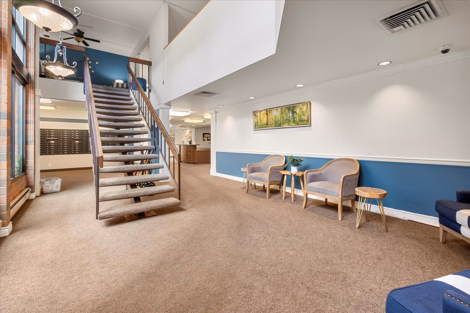 Interior view of a senior living facility lobby with a carpeted staircase in the center, two upholstered chairs with a small wooden table and plant between them on the right, and a reception desk in the background. The walls are painted white with a blue accent panel, and there is a framed nature painting above the chairs. Large windows and hanging light fixtures are visible on the left side.
