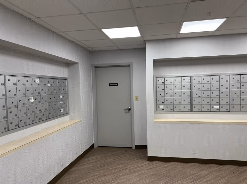 Interior hallway area with two sets of metal mailboxes mounted on opposite walls and a closed gray door labeled 'Employees Only' in the center. The walls have light-colored patterned wallpaper, and the floor is covered with wood-like vinyl flooring. The ceiling has white acoustic tiles with recessed lighting.