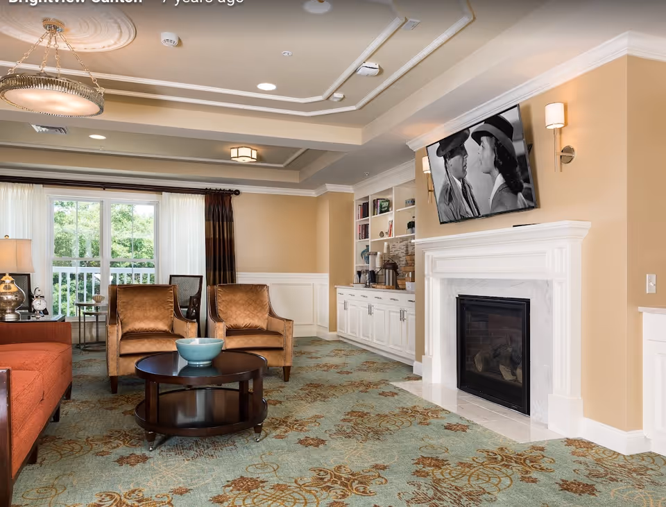 A cozy senior living room with two brown armchairs, a round wooden coffee table with a blue bowl, an orange sofa, a fireplace with a mounted TV showing a black and white movie, built-in white cabinets with a coffee station, and large windows with curtains letting in natural light.