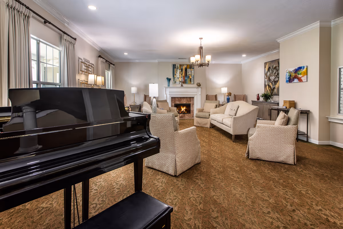 A spacious living room with a black grand piano in the foreground, beige upholstered chairs and sofas arranged around a lit fireplace, soft lighting from lamps and a chandelier, and colorful artwork on the walls.