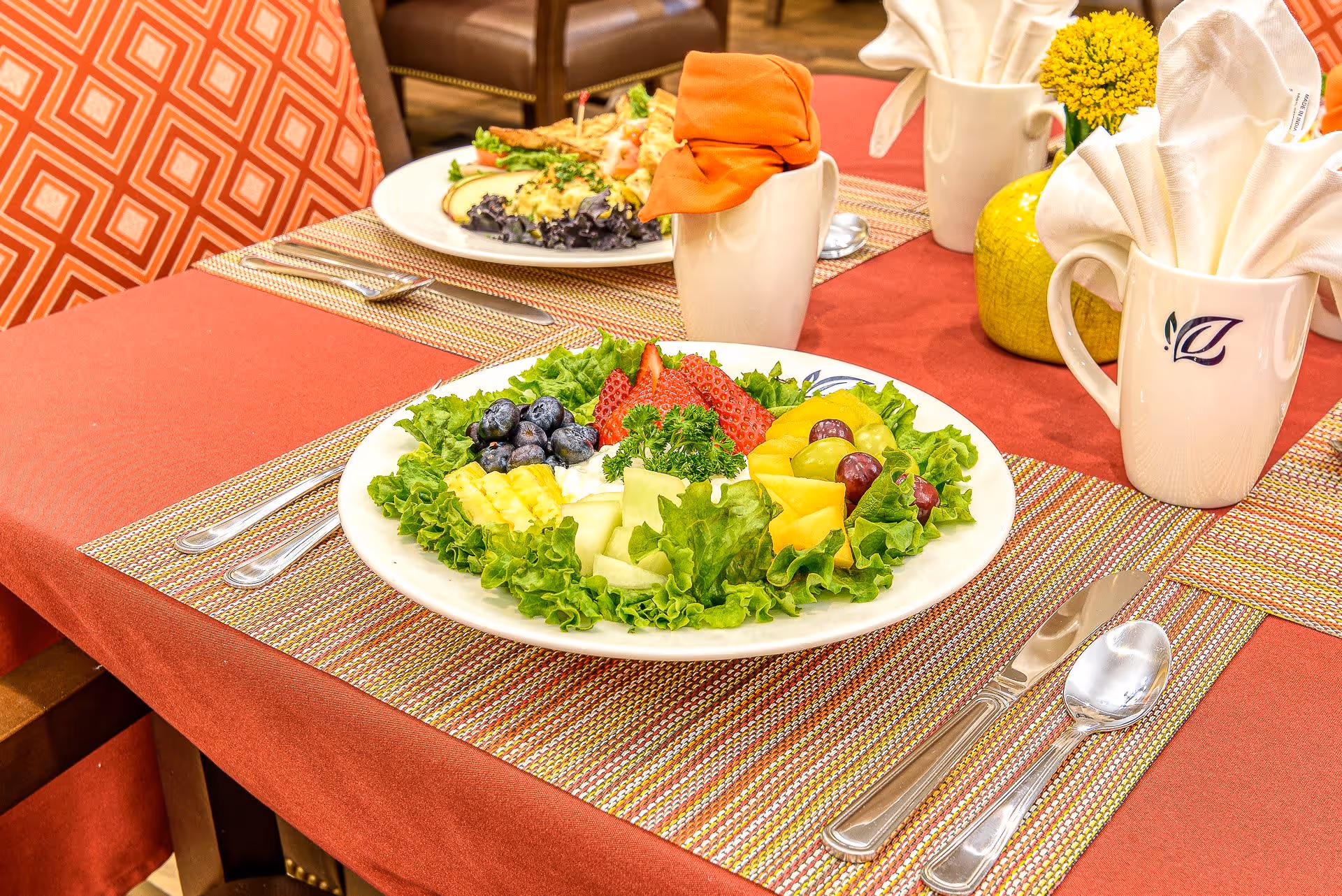 A dining table set with a plate of fresh fruit salad including strawberries, blueberries, grapes, pineapple, cantaloupe, and melon on a bed of lettuce. The table has a red tablecloth with a woven placemat, silverware, and white mugs with folded napkins. There is a yellow flower in a small vase on the table and an orange napkin in one of the mugs.